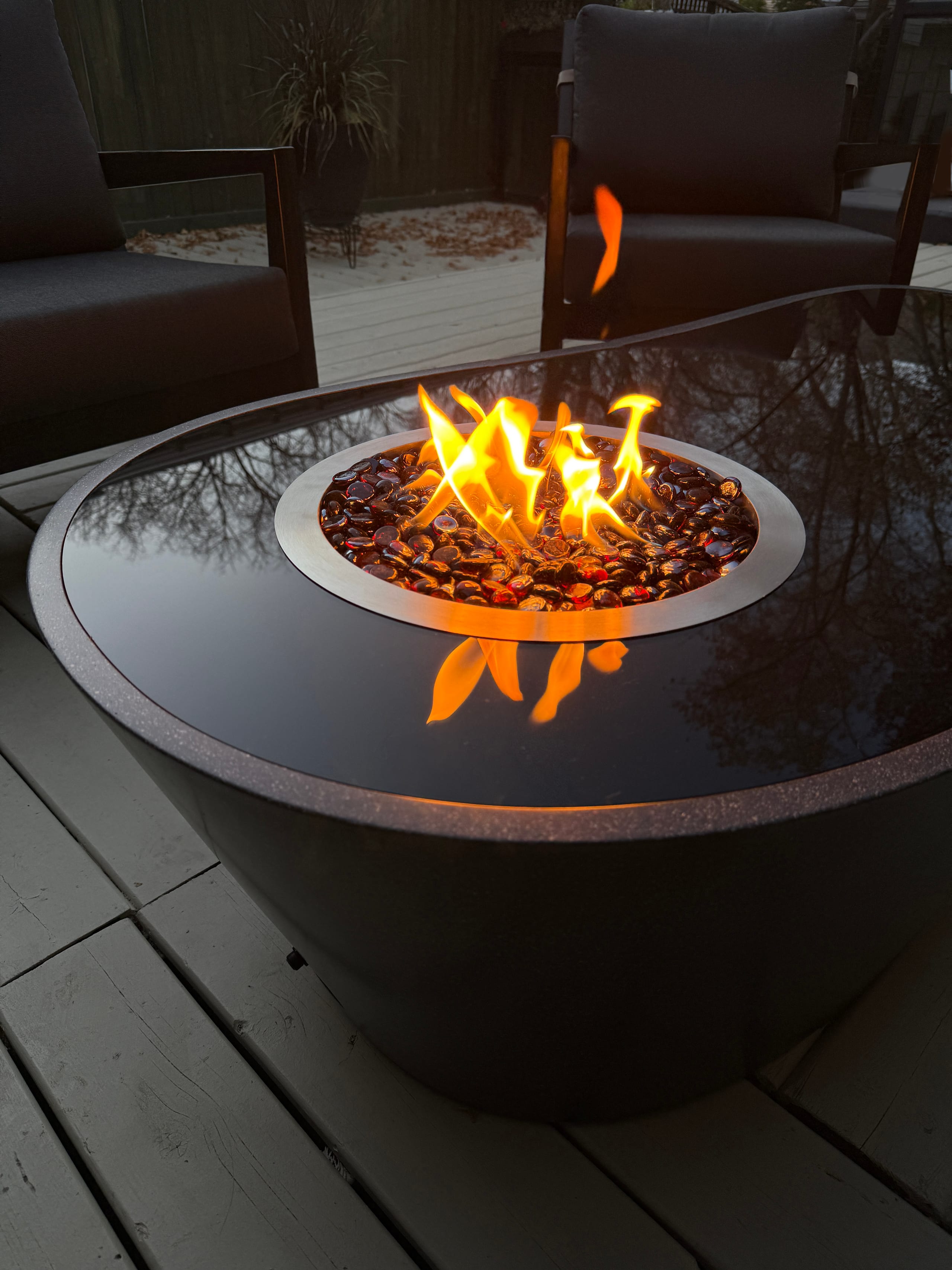 Round dark-topped gas fire pit with orange flames over glass fire beads on a light wooden deck, with dark patio chairs behind it at dusk