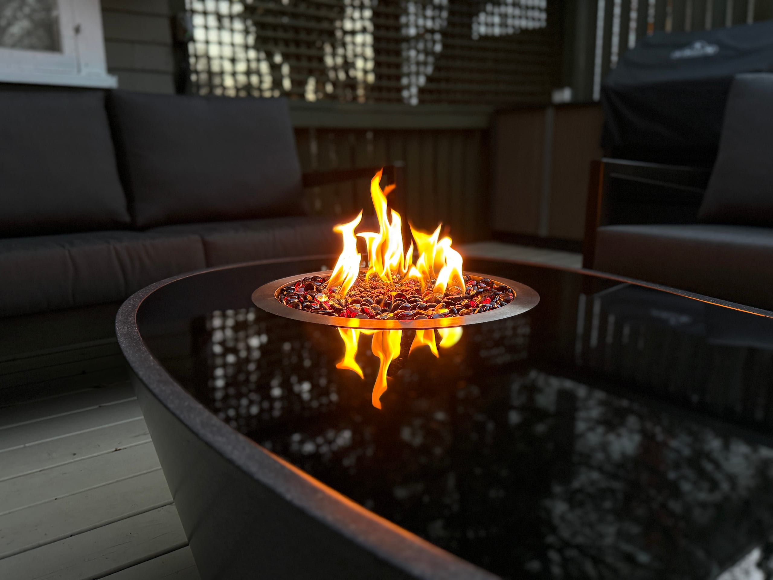 Round outdoor fire pit with colourful glass stones and bright flames, surrounded by charcoal patio lounge chairs on a composite deck at dusk